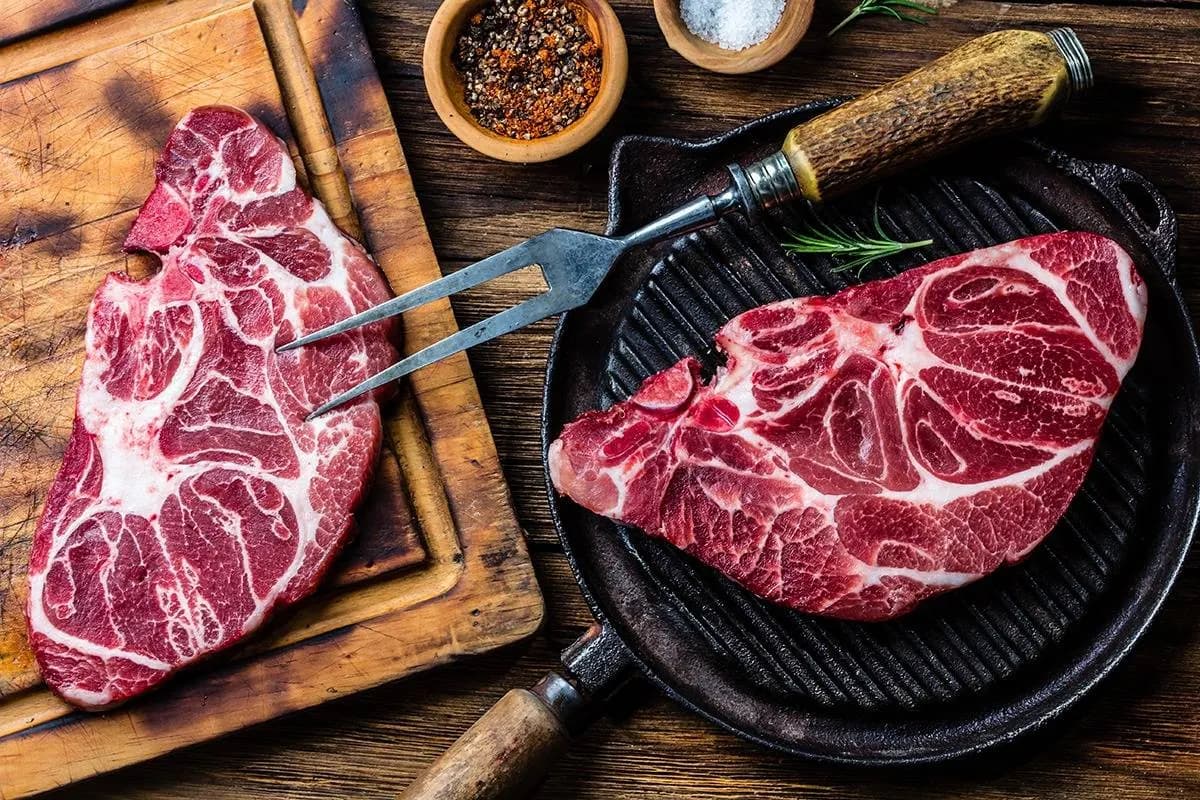 Thick, well-marbled raw beef on a wooden board beside a cast iron pan, with bowls of salt and cracked pepper.