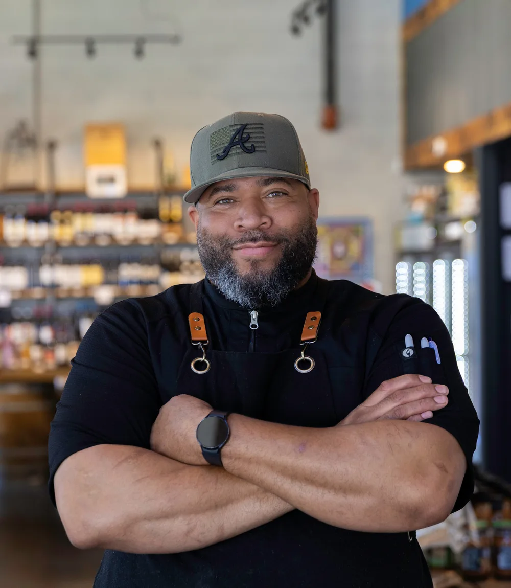 Chef Antwan Cooper, known as Chef De, in a black chef's jacket and apron with his arms crossed, in front of the market shelves at Frazie's Meat & Market.