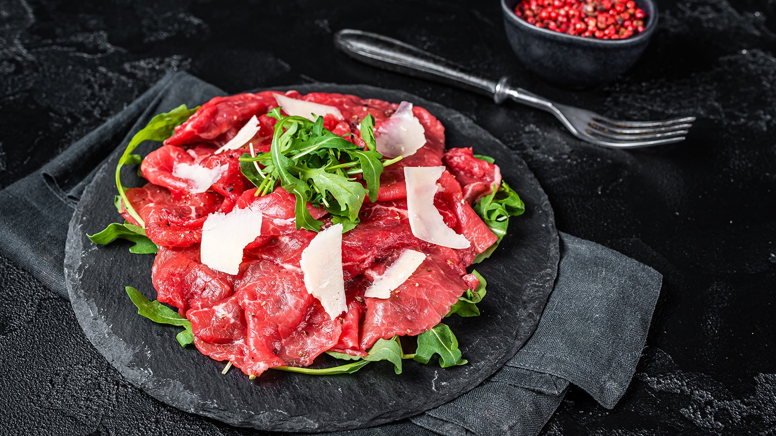 An artful presentation of beef Carpaccio, garnished with fresh arugula leaves and shavings of Parmesan cheese. The thinly sliced, ruby-hued raw beef arranged in a circle contrasts with the bright green of the arugula and the white Parmesan. The image evokes an enticing promise of a delightful blend of textures and flavors.