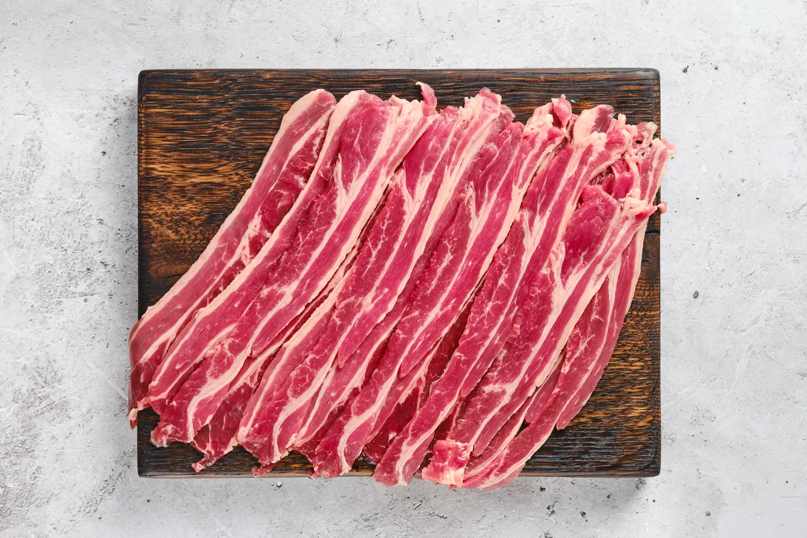 Raw beef bacon sliced into thick strips and laid out on a wooden cutting board, showing the lean and fat layers of the navel cut.